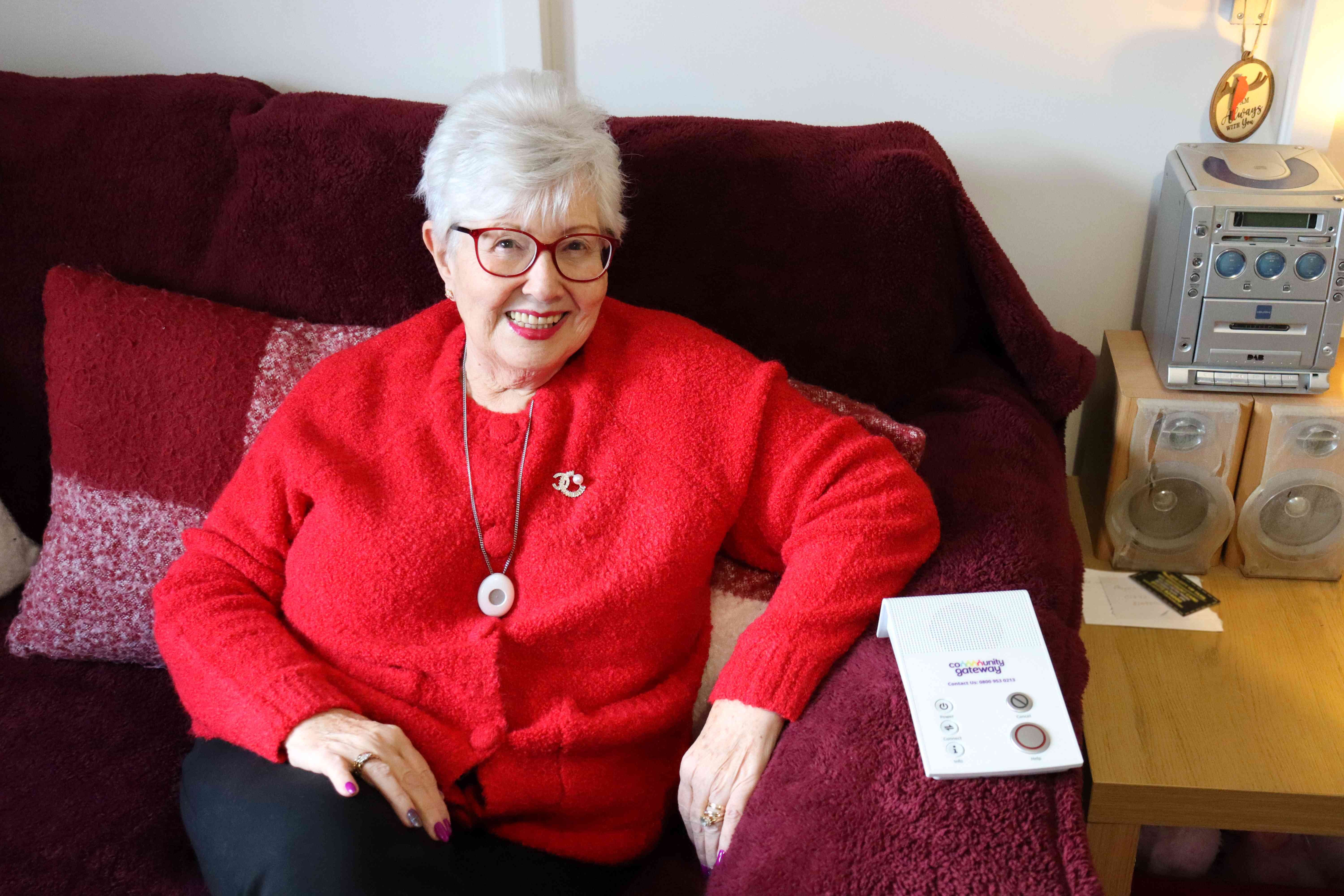Lady with short white hair in a red cardigan and red glasses smiling at the camera with a lifeline unit text to her arm