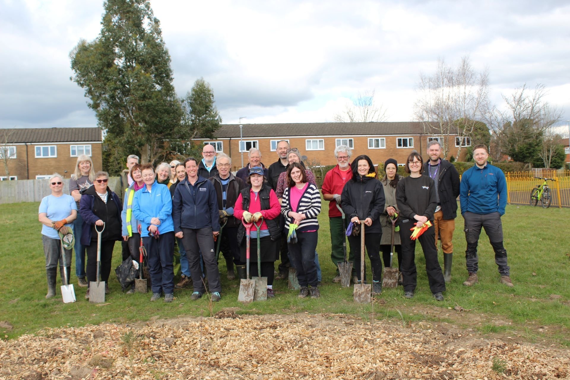 A large group of people stood with shovels in a green space, having planted a micro-wood in Ingol