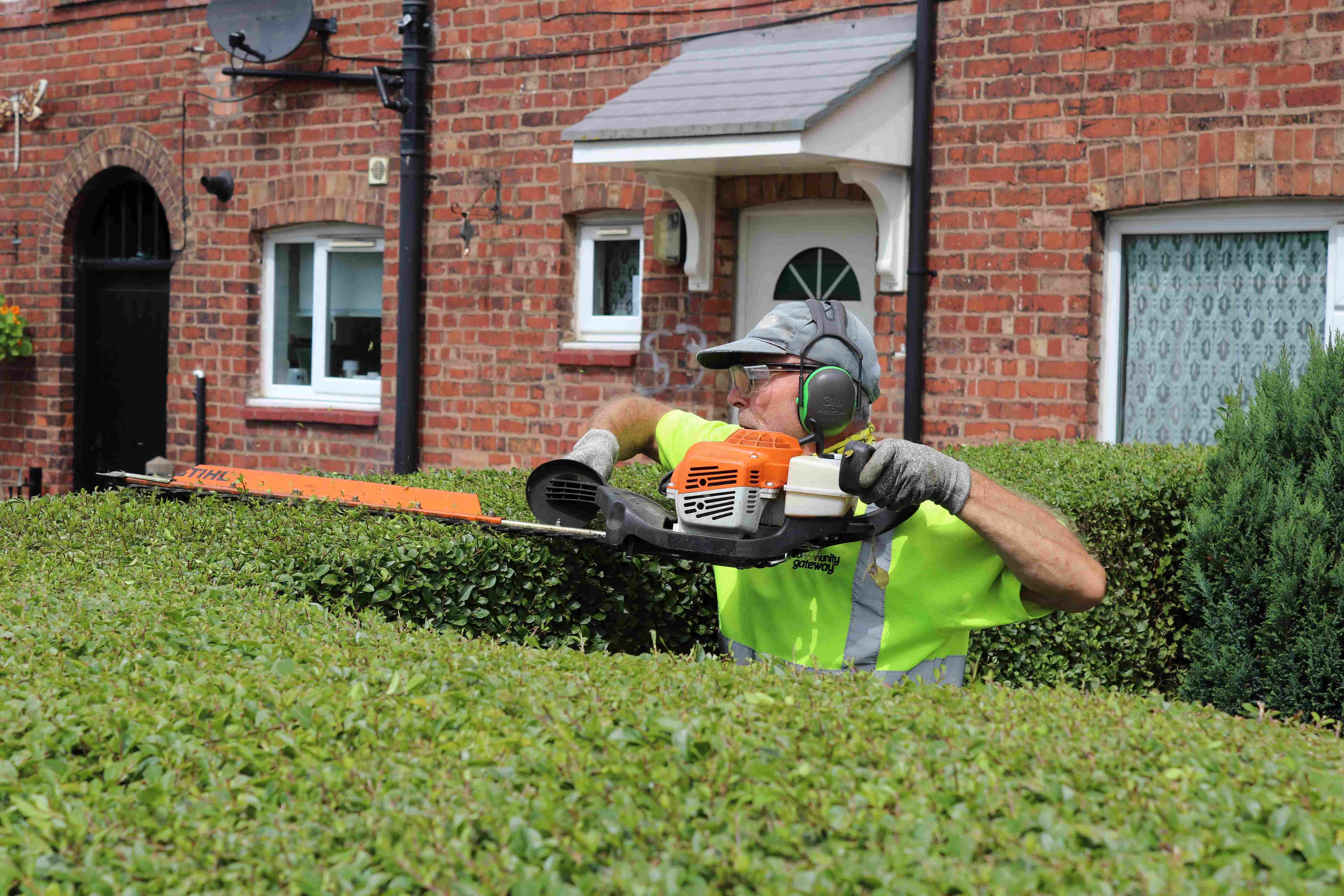 A person wearing high vis with headphones on and a hedge trimmer, working on a building outside a red brick house